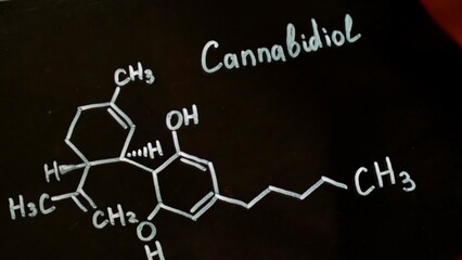 Close-up drawing of the chemical formula of cannabidiol on a black background. A scientist chemist writes in white