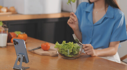 A young woman with a beautiful face in a blue shirt with long hair eating fruit sitting inside the kitchen at home with a laptop and notebook for relaxation, Concept Vacation.