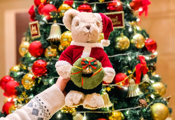 A hand holding a Christmas teddy bear in festive outfit holding an xmas wreath in front of Christmas tree