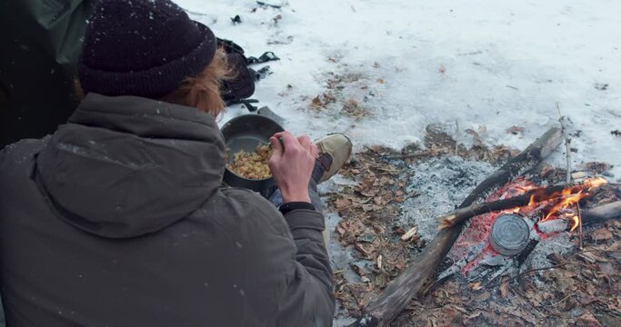 hungry man eating food from the pot The tourist camping, outdoors, winter slow motion traveler has rest, take break Top view Winter survival concept. bushcraft, lifestyle