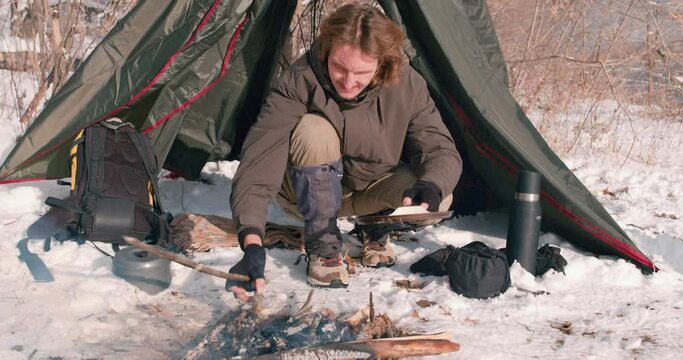 traveler sitting next to the tent going to cook food at campsite in winter Slow motion. A young caucasian man prepares a dish. Winter survival concept. bushcraft, man with frying pan over campfire