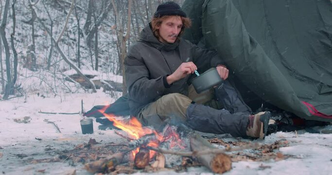 man sitting at campfire cooking breakfast at campsite on snowy day in winter Tourist stiring mixing porrage Hicker eats food on hike from pot Camping utensils Survival in wildlife Lifestyle, hobby