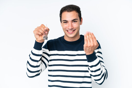 Young caucasian man holding home keys isolated on white background inviting to come with hand. Happy that you came