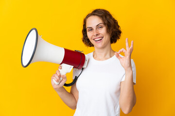 Young English woman isolated on yellow background holding a megaphone and showing ok sign with...