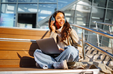 Happy woman in stylish clothes with laptop and headphones in the city works. Young woman listening to music, video conference or podcast. Lifestyle. Freelancing concept, online education.