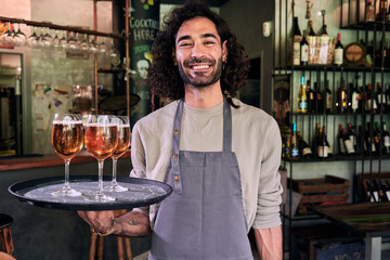 Cheerful waiter looking at camera while holding a tray with beers at place of work. Entrepreneur young male at new restaurant.