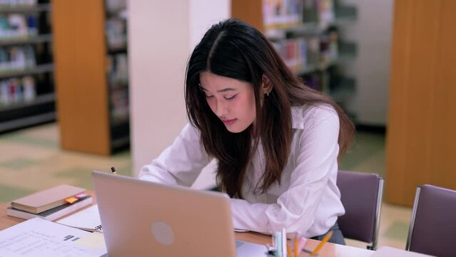 Concentrated teen Asian female student in casual clothes working hard on Laptop for a research, Writes Notes for the Paper in University Library