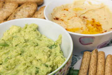 A close-up shot of a bowl of guacamole dip, made with fresh avocado, onion, and cilantro. The background is blurred, but you can see a bowl of hummus, whole wheat toast and bread sticks.