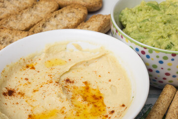 A close-up shot of a bowl of hummus dip, made with chickpeas, tahini, garlic and lemon. The background is blurred, but you can see a bowl of guacamole, whole wheat toast and bread sticks.