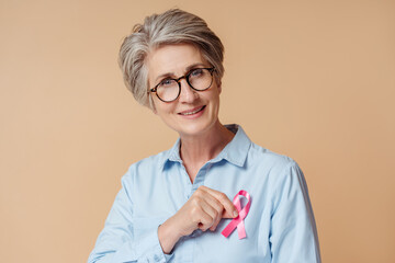 Portrait of smiling confident senior woman holding  pink ribbon isolated on beige background. Health care, support, prevention. Breast cancer awareness month concept 