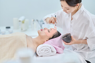 아시아 한국의 젊은 여성이 스파 또는 피부관리전문점 또는 피부과 병원에서 분홍색 두건을 쓰고 침대에 누워 베이지색 대형 타올을 덮고 하얀색 셔츠를 입은 전문관리사가 실리콘붓으로 모델링팩을 얼굴에 바르고 있다. 
