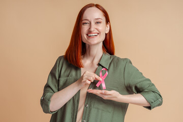 Beautiful smiling confident red haired woman holding breast cancer pink ribbon isolated on beige...