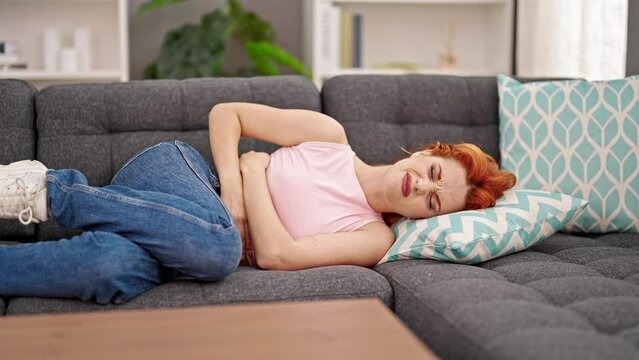 Young redhead woman suffering for menstrual pain lying on sofa at home