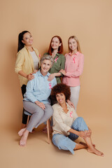 Group of smiling multiethnic women wearing breast cancer pink ribbon support each other celebrate recovery isolated on beige background. Health care, prevention. Breast cancer awareness month