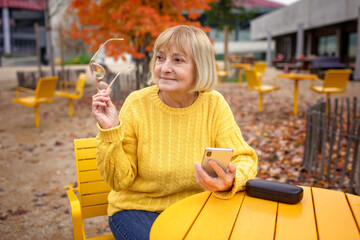 Thoughtful woman in her late sixties sits in the street cafe in autumn, surrounded by fall colors. She holds eyeglasses and smartphone. Digital era, a tech-savvy senior and modern technology