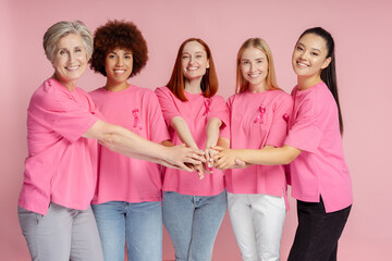 Group of smiling multiracial women wearing t shirts with breast cancer pink ribbon holding hands together isolated on pink background. Health care, support. Breast cancer awareness month concept