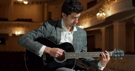 Guy in elegant stylish gray suit plays the guitar sitting on chair on the stage. Recital of a young musician. american brunette Guitar player performing solo sound at concert Hobby Interest Profession