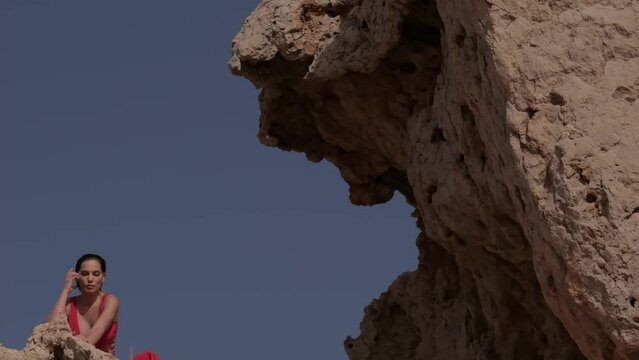 A young woman in a red dress is sitting on a rock in the middle of the desert against a blue sky.