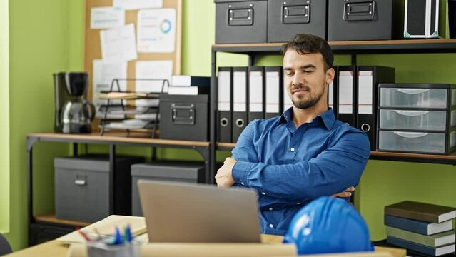Young hispanic man architect using laptop sitting on table with arms crossed gesture at office