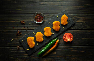 Chicken nuggets on a serving board with spices cayenne peppers and tomatoes. Fast food concept on black table