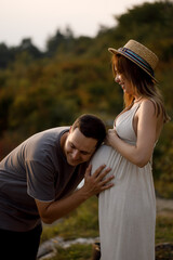 A young couple in love are standing against the background of the sunset, looking at each other, smiling. A woman holds a pregnant belly in her hands.