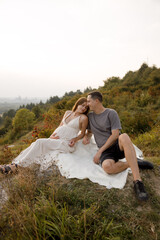 A young couple in love are sitting on a white blanket, against the background of the sunset, looking at each other, smiling. A woman holds a pregnant belly in her hands.