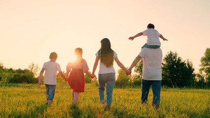 Happy family walks towards sun on green grass.Mom dad children play in nature. Parents with children, holding hands, play in park. Happy Family walk. People in park, holding hands, walk across field