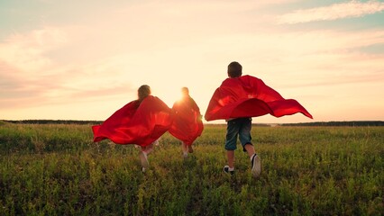 Happy girls, boy kids play superheroes they run across green field in red cloak, cloak flutters in...