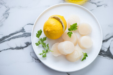 White plate with raw fresh scallops, lemon and parsley, horizontal shot on a grey marble background, elevated view