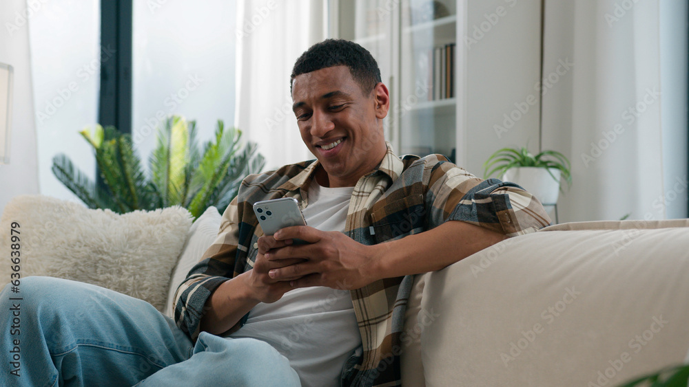 Wall mural african american guy ethnic happy smiling man relaxing on comfortable couch sofa at home using mobil