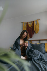 Fototapeta premium Young adult woman with smartphone sitting in bed in morning, wrapped in blanket