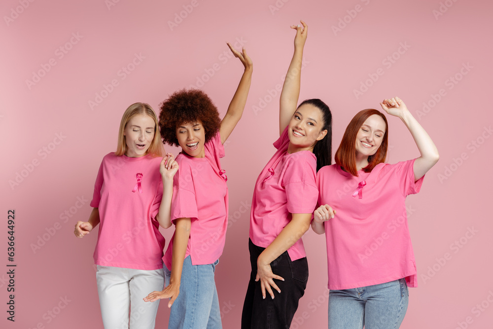 Wall mural Group of smiling multiethnic women wearing t shirts with pink ribbon dancing, celebration isolated on pink background. Breast cancer awareness month