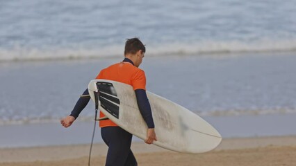 A man in orange t-shirt walking to the shore holding a surfboard