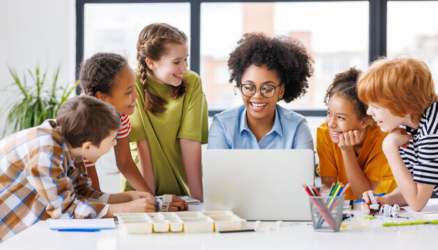Cheerful  schoolkids  with teacher looking at laptop screen and study the scheme of assembly of robots during collective school work    .