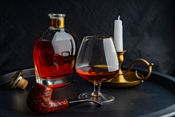 A glass of cognac against the background of a bottle of cognac of a white candle in a bronze candlestick, on a dark tray on a dark background and a pipe for smoking in the foreground