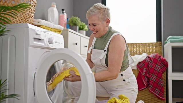 Middle age grey-haired woman smiling confident washing clothes doing thumb up gesture at laundry room