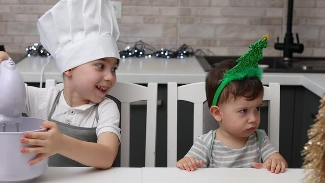 siblings boys doing with granny gingerbread christmas house.preschooler child wearing toque chef  hat,apron,mixing cream,licking from wires beaters.toddler with tree headband,modern kitchen 4k
