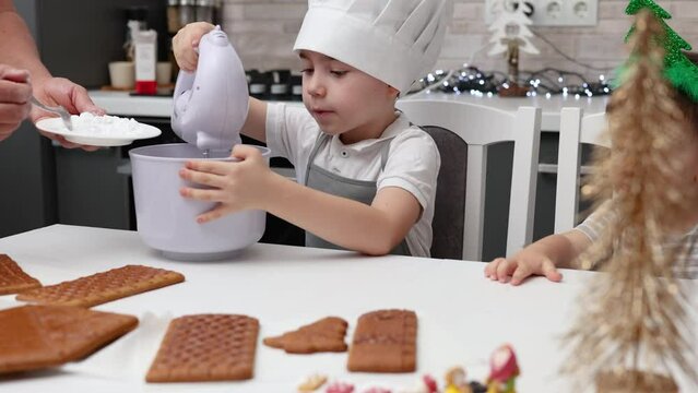 siblings boys doing with granny gingerbread christmas house.preschooler child wearing toque chef  hat,apron,mixing cream,licking from wires beaters.toddler with tree headband,modern kitchen 4k
