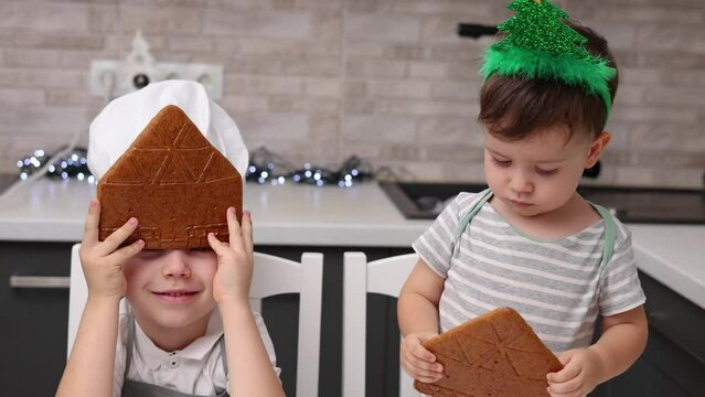 siblings boys doing with granny gingerbread christmas house.preschooler child wearing toque chef  hat,apron,mixing cream,licking from wires beaters.toddler with tree headband,modern kitchen 4k