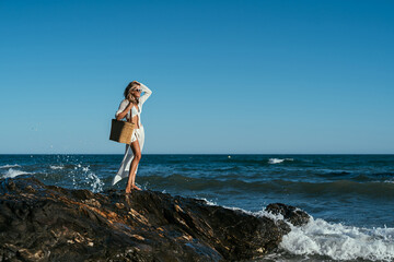 A beautiful girl in white glasses and white clothes with a bag in her hands stands on the stones on the seashore