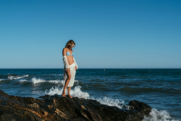 A beautiful girl in white glasses and white clothes sits on the rocks on the seashore