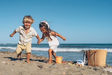 a little blond boy and a little brunette girl holding hands looking at the sea on the beach standing on the sand in front of the water on a summer sunny day