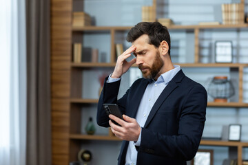 Serious thinking businessman standing near window at workplace, senior boss received online notification on phone with bad news, man in suit reading disappointed, using app on smartphone.