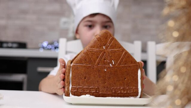 siblings boys doing with granny gingerbread christmas house.preschooler child wearing toque chef  hat,apron,mixing cream,licking from wires beaters.toddler with tree headband,modern kitchen 4k
