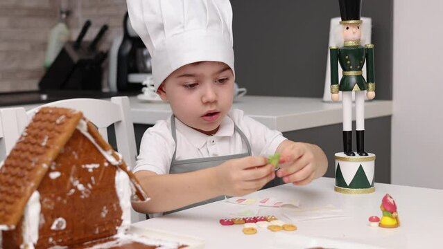 siblings boys doing with granny gingerbread christmas house.preschooler child wearing toque chef  hat,apron,mixing cream,licking from wires beaters.toddler with tree headband,modern kitchen 4k
