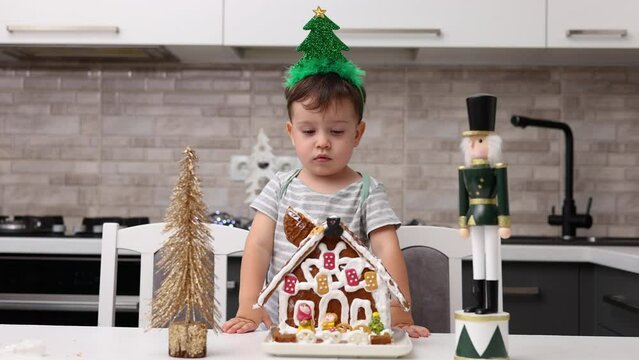 siblings boys doing with granny gingerbread christmas house.preschooler child wearing toque chef  hat,apron,mixing cream,licking from wires beaters.toddler with tree headband,modern kitchen 4k