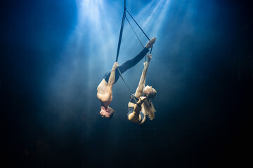 An aerial straps duo performs dangerous tricks mid-air, illuminated by strong blue and white lights against a dark background, clad in minimal athletic attire, showcasing a blend of power and grace