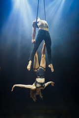 An aerial straps duo performs dangerous tricks mid-air, illuminated by strong blue and white lights...