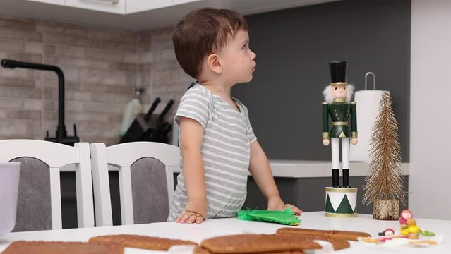 siblings boys doing with granny gingerbread christmas house.preschooler child wearing toque chef  hat,apron,mixing cream,licking from wires beaters.toddler with tree headband,modern kitchen 4k