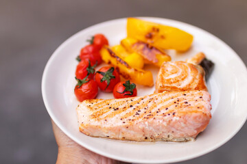 Fototapeta na wymiar Hand holding plate of salmon fillet steak with cherry tomatoes and bell pepper.